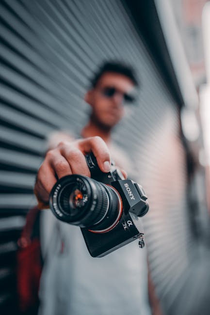 A person extends a Canon camera towards the viewer in an outdoor setting.